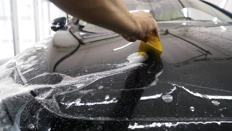 A person using a yellow squeegee to wash soap suds off the hood of a black car with sleek car wraps.