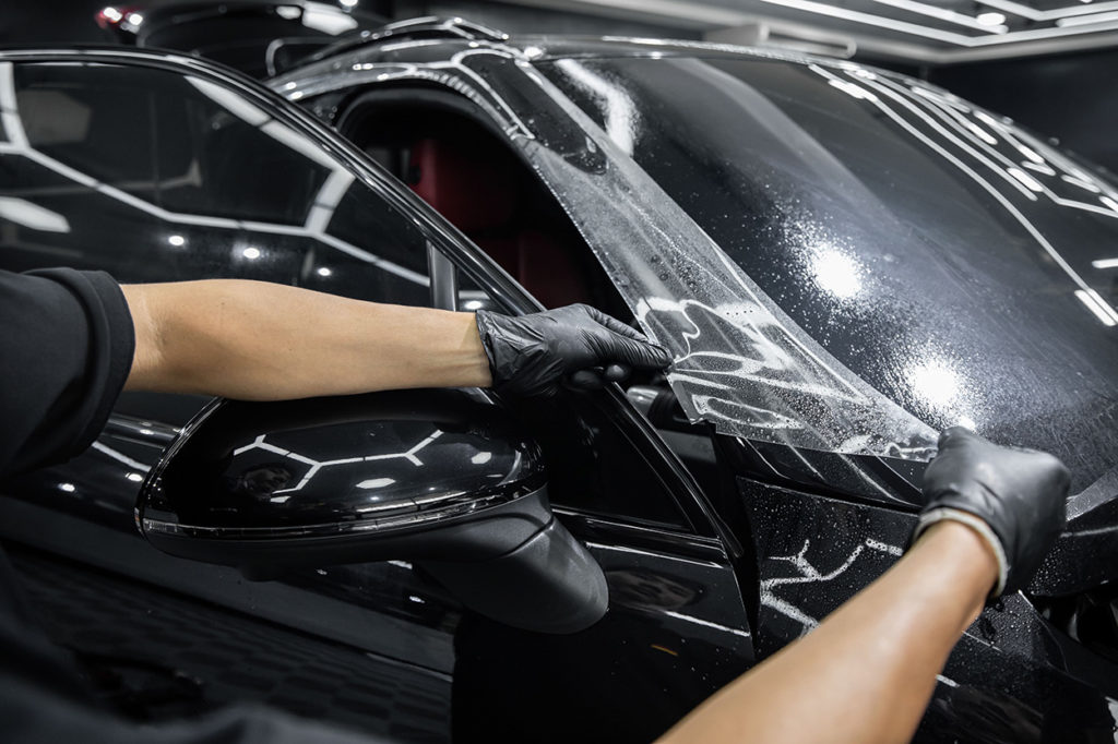 In a well-lit detailing shop, a person carefully applies protective film to the black car's hood and fender, ensuring the car wrap fits perfectly.