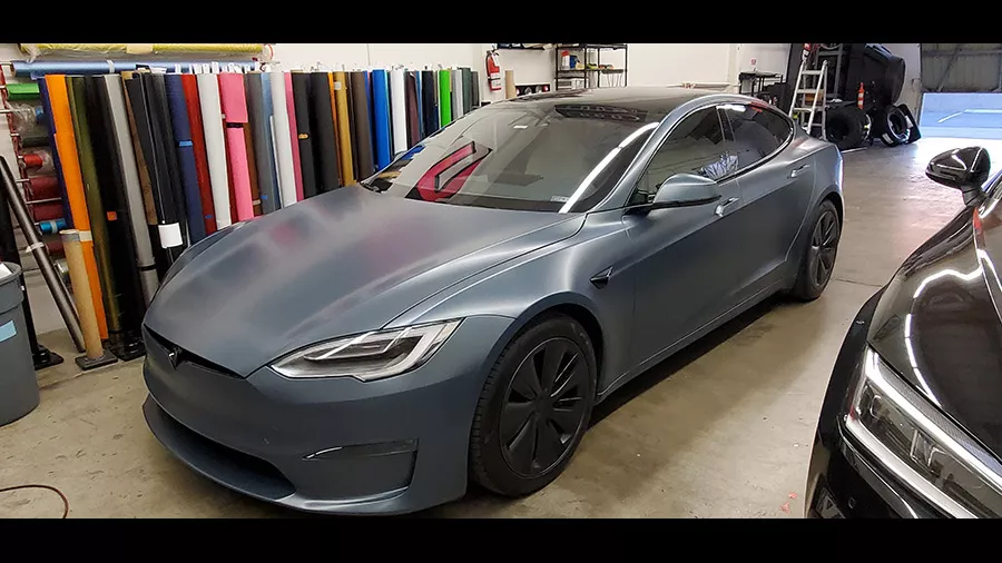 A matte blue Tesla Model S parked indoors near rolls of colored vinyl wrap and a black car.