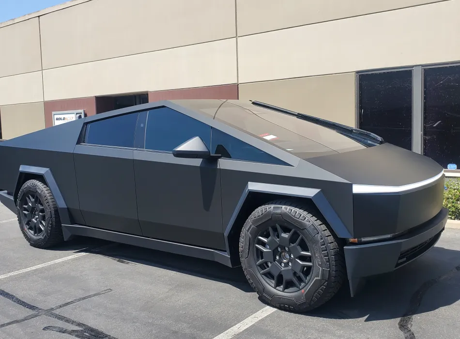 A matte black Tesla Cybertruck is parked in a lot beside a beige building on a sunny day.