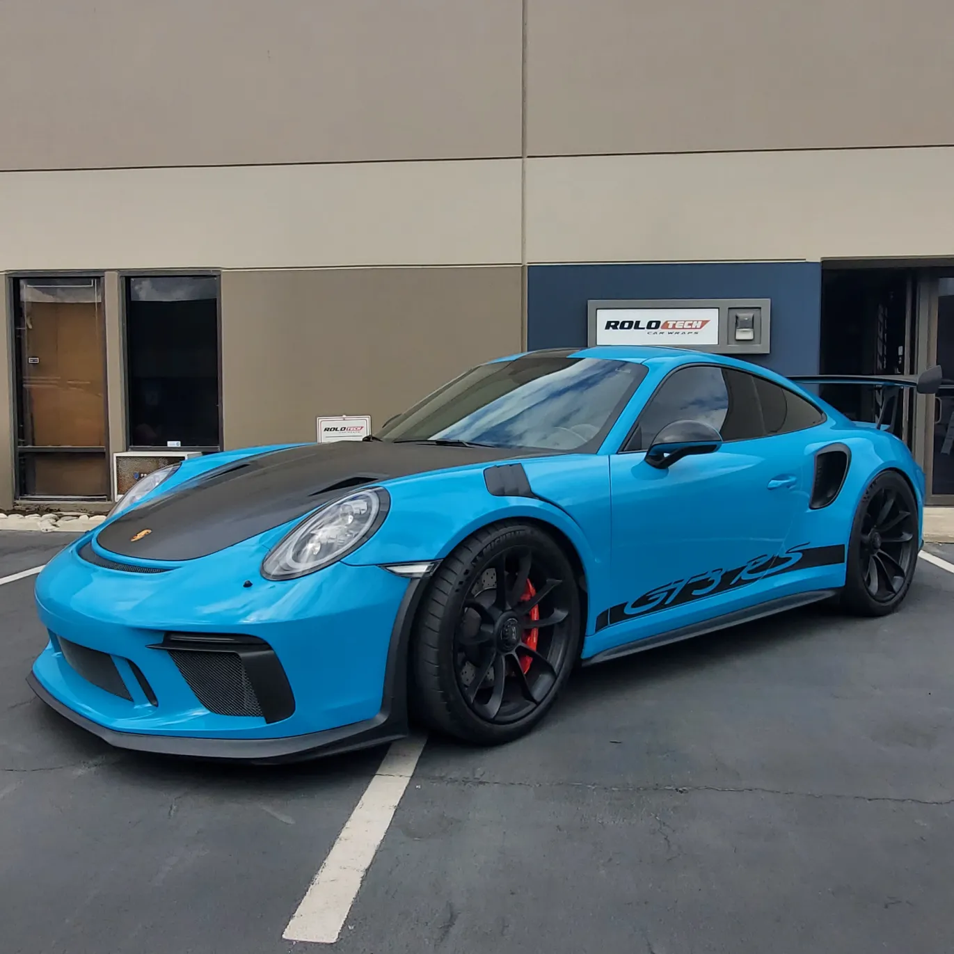 A blue Porsche sports car with black accents parked in front of a building with tan and gray walls.