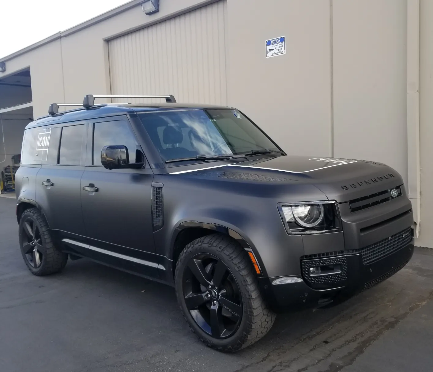 Matte black Land Rover Defender SUV parked beside a beige industrial building.