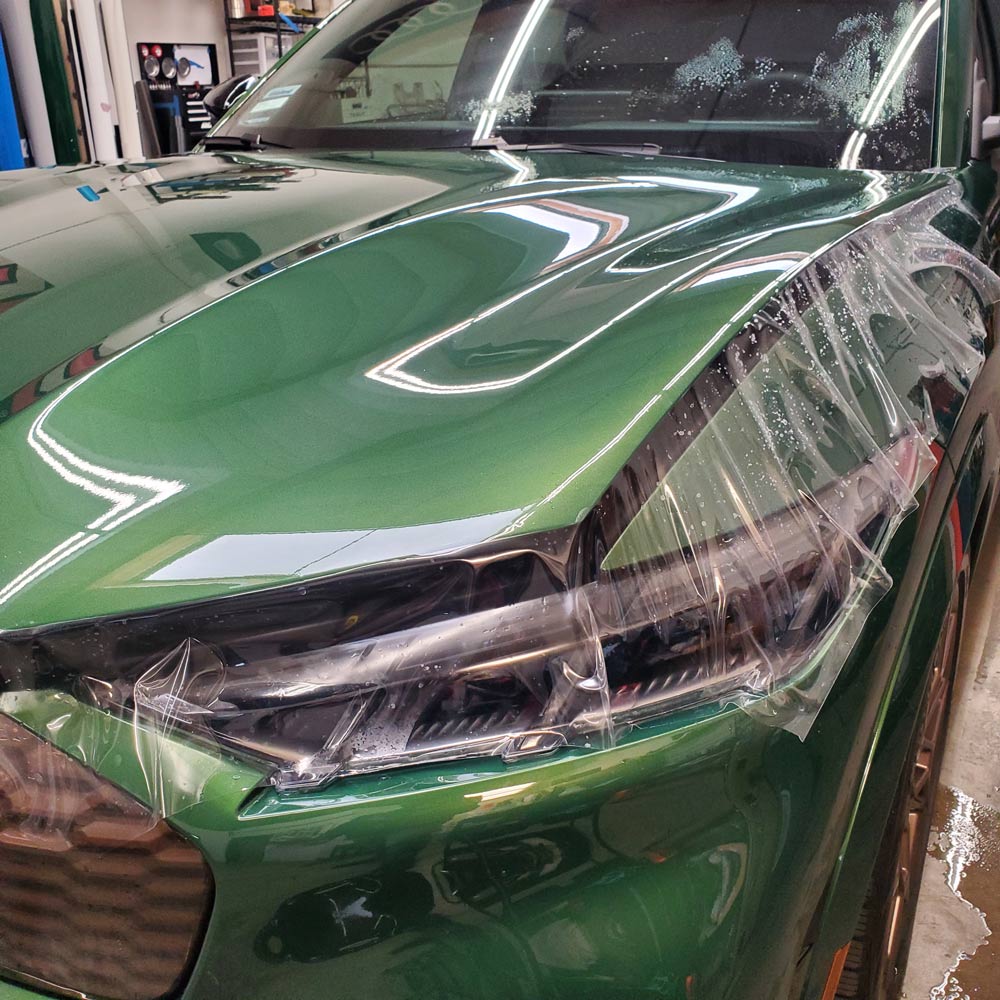 20241205_102404 Clear protective film being applied to the front hood and headlight of a shiny green car in a workshop.