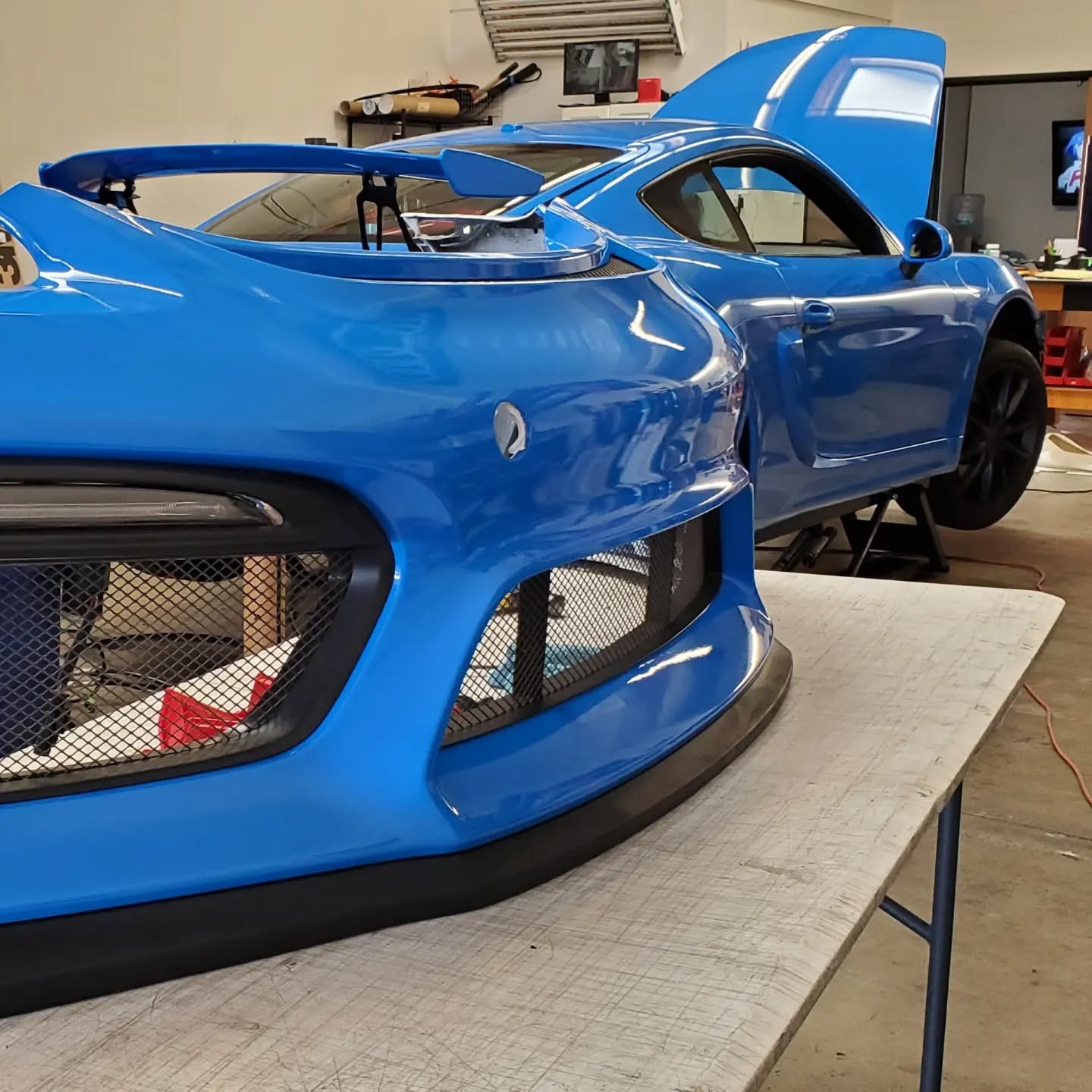 A bright blue sports car in a garage with its hood open and front bumper removed, resting on a table.