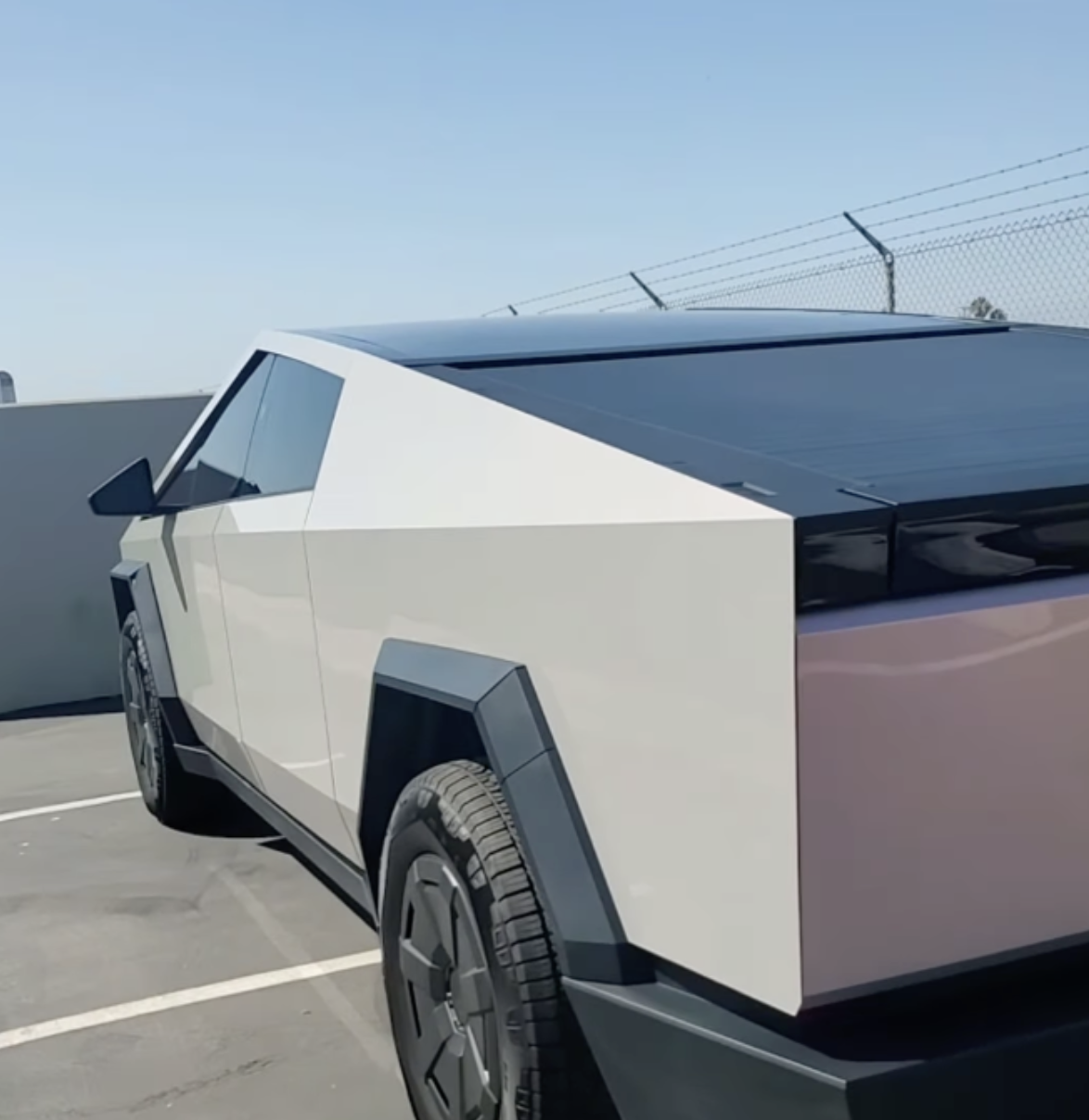 Color PPF vs Vinyl Wrap in Orange County A silver Tesla Cybertruck is parked in an outdoor parking lot near a fence on a sunny day.