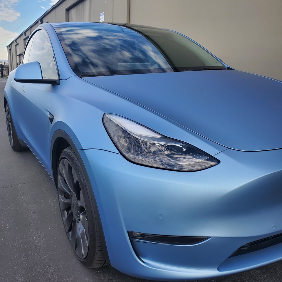 A blue Tesla car parked outside next to a beige building under a partly cloudy sky.