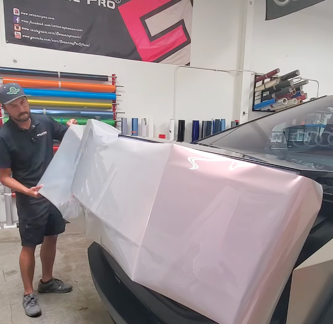 A man applies a large sheet of pink car wrap to the front of a vehicle in a workshop.